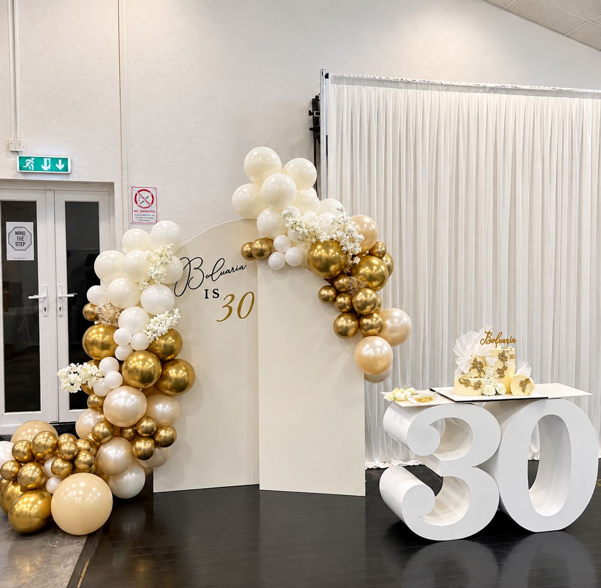 Ivory and gold balloon garland arching over two backdrops, with a cake table set on giant gold 30 numbers